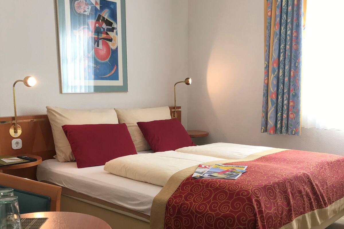 Habitación de hotel con cama, dos lámparas de noche, una mesa con vasos, un cuadro en la pared y una ventana con cortinas.