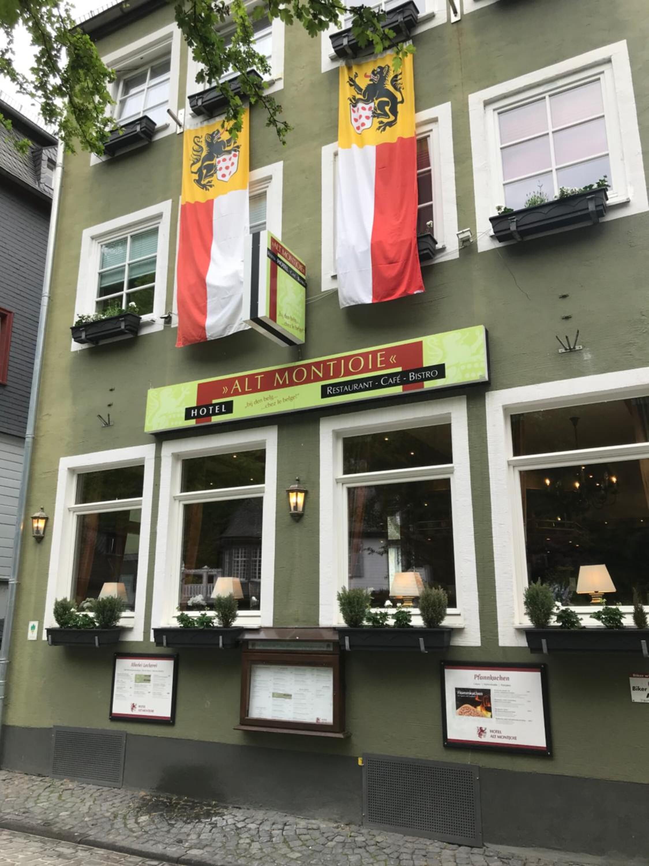 Een gebouw met een groene gevel, ramen met witte kozijnen en twee vlaggen. Er staan borden met 'Alt Montjoie' op, wat aangeeft dat het een restaurant, café en bistro is.