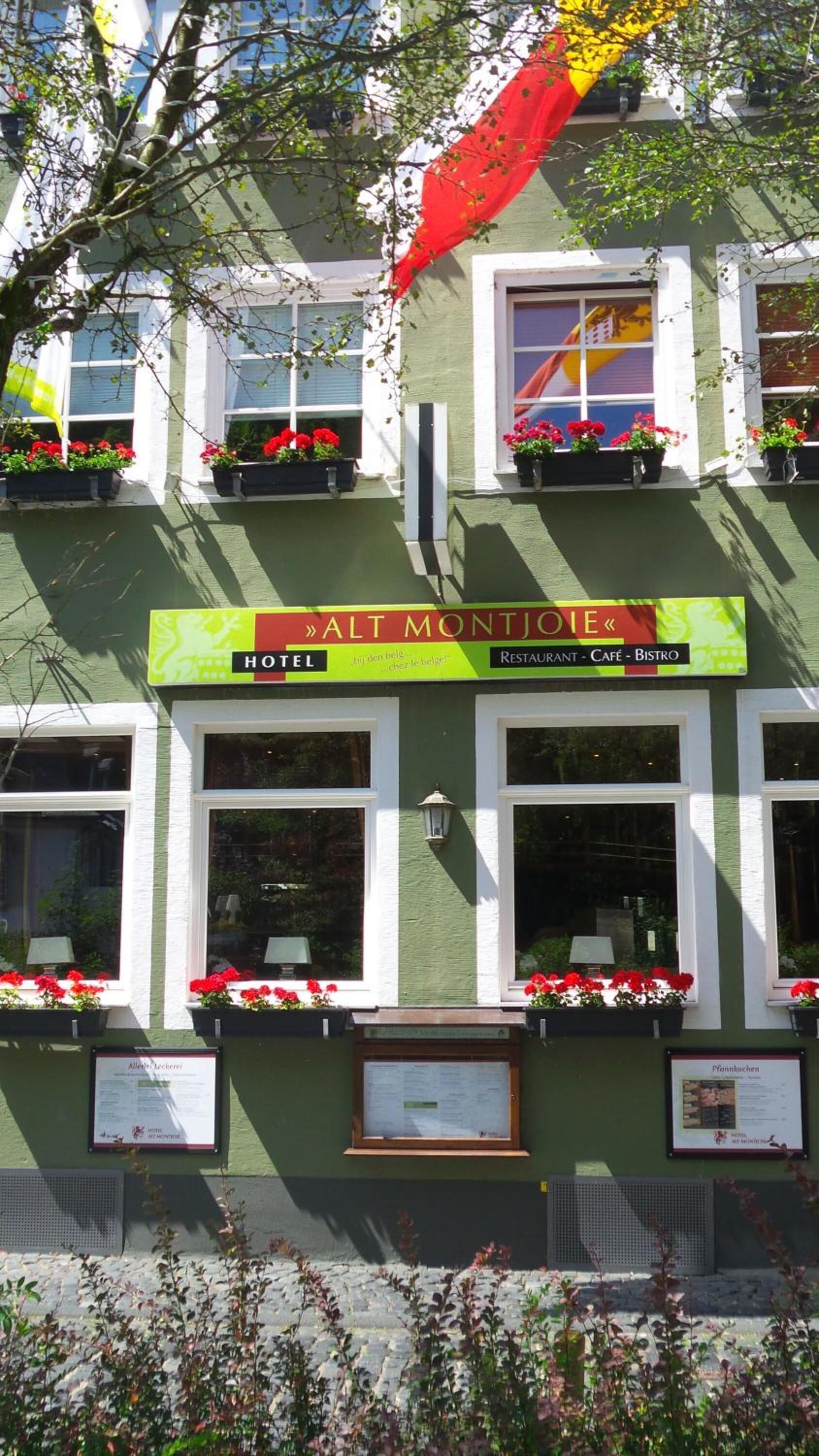 A building with a green facade, multiple windows, and flower boxes with red flowers. There is a sign above the windows that reads "Alt Montjolie" and indicates it is a hotel, restaurant, café, and bistro.
