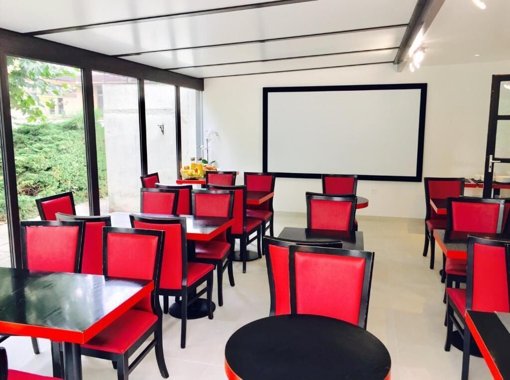 A conference room with red chairs and tables, a projector screen, and a view of greenery outside.