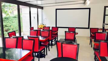 A conference room with red chairs and tables, a projector screen, and a view of greenery outside.