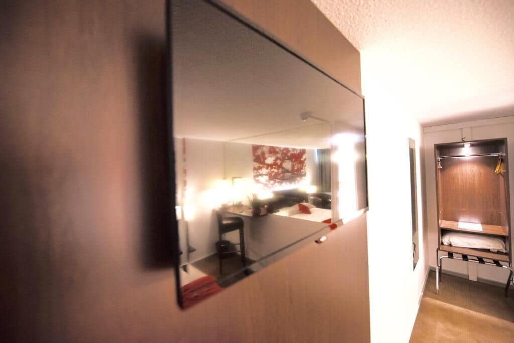 Habitación de hotel con un espejo grande que refleja el interior, un armario de madera y una pequeña zona de estar con una silla y una mesa.