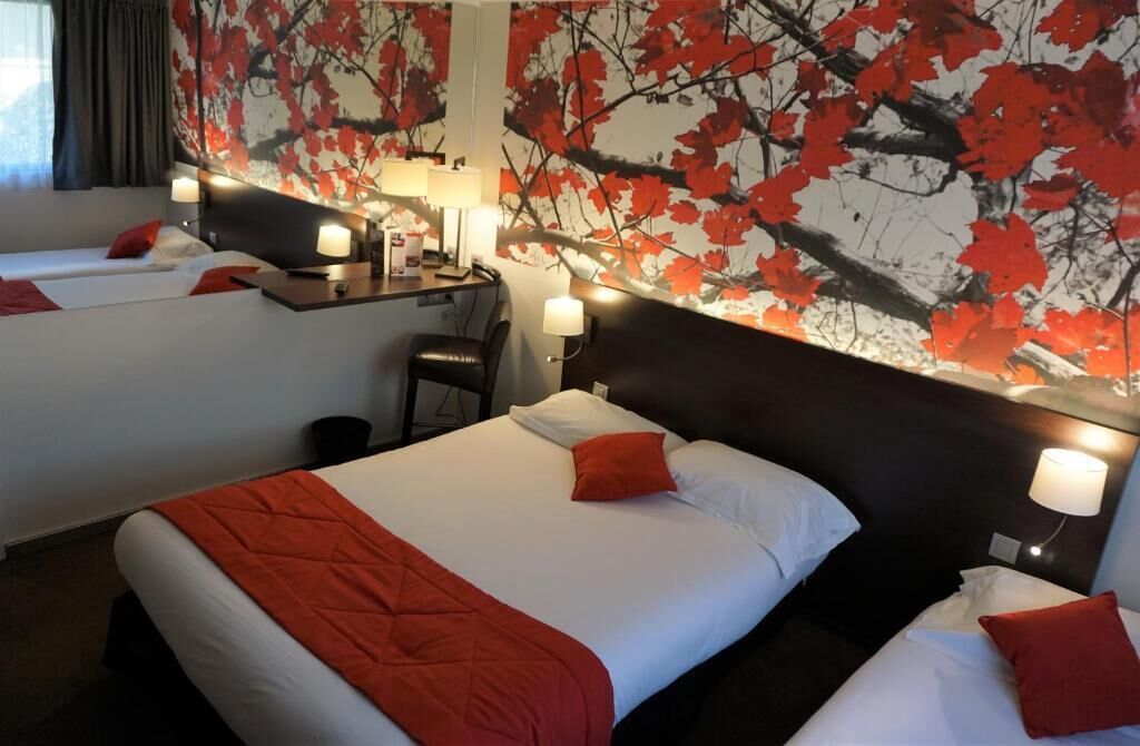 A hotel room with two beds, each with a red pillow and white bedding. There is a desk with a lamp and a chair. The wall features a red and black leaf pattern.