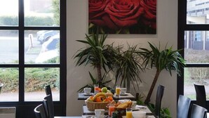 A dining area with a table set for breakfast, featuring a fruit bowl, orange juice, and a painting of red roses on the wall. - Hôtel Pavillon des Gâtines (Plaisir)