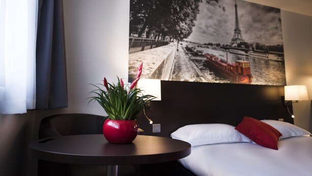 A hotel room with a bed, a small round table with a red pot, and a wall-mounted lamp. - Hôtel Pavillon des Gâtines (Plaisir)