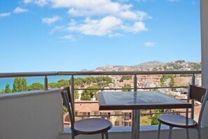 Ein Balkon mit Tisch und zwei Stühlen, mit Blick auf eine Küstenstadt mit Häusern und Bäumen.