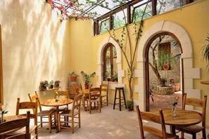 Une terrasse ensoleillée, dotée de tables et de chaises en bois, entourée de plantes en pot et offrant une vue sur un jardin à travers une fenêtre en arc de cercle.