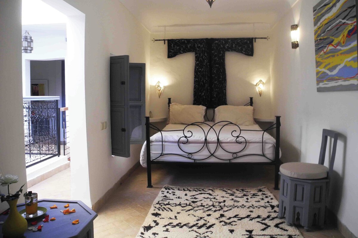 Un dormitorio con cama, mesita de noche, silla, ventana y puerta.