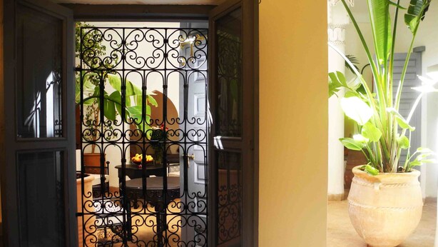 An open door with a wrought iron gate, leading to a patio with a table and chairs, and a potted plant on the right. - Riad Linda (Marrakech)
