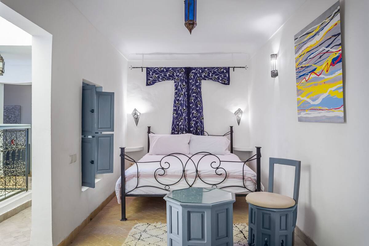 A bedroom with a bed, a chair, a small table, a wardrobe, and a painting on the wall.