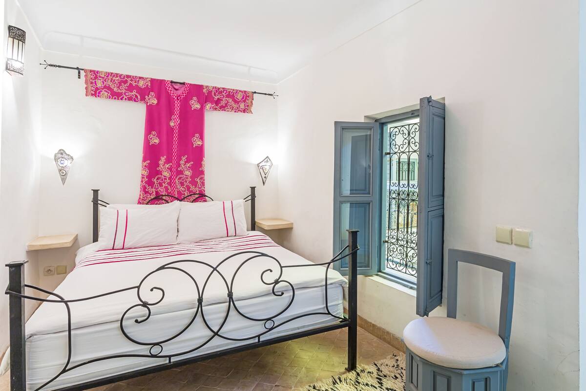 A bed with a metal frame, a window with blue shutters, and a chair with a cushion.