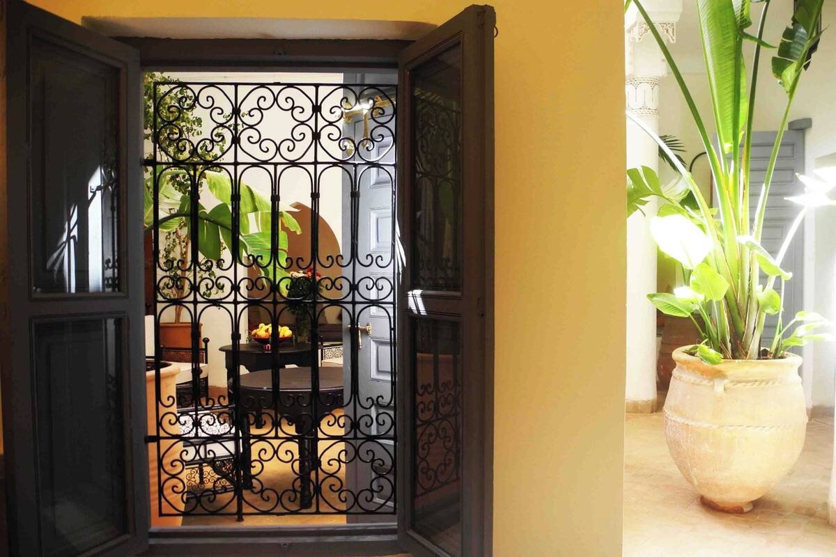 An open door with a wrought iron gate, leading to a patio with a table and chairs, and a potted plant on the right.