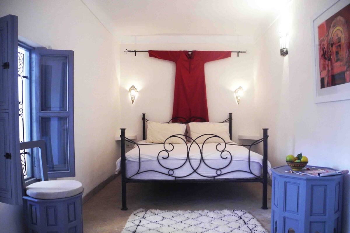 A bedroom with a bed, a nightstand, a stool, a red curtain, and a wall-mounted light fixture.
