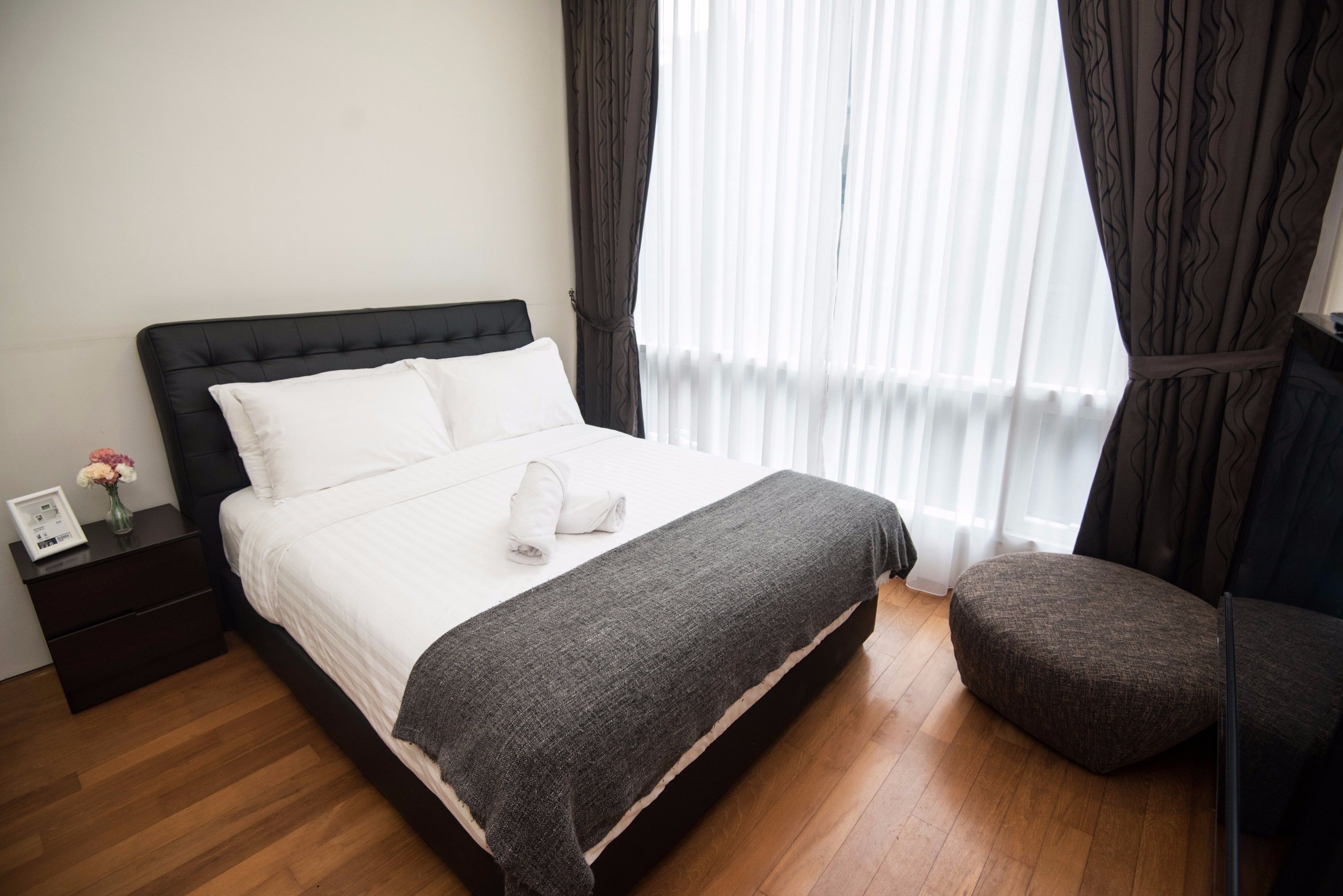 A neatly made bed with a white duvet and a grey throw blanket, a small bedside table with a vase of flowers, and a wooden floor.