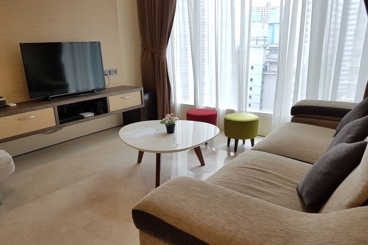 Una sala de estar moderna con una televisión de pantalla plana, una mesa de centro redonda, un sofá color beige y dos otomanas.