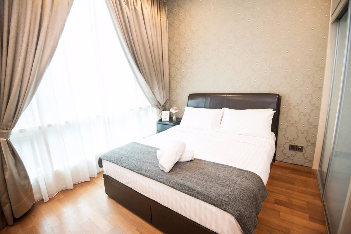 Una cama bien hecha con sábanas blancas y una manta gris, suelo de madera y una pared con un diseño discreto.