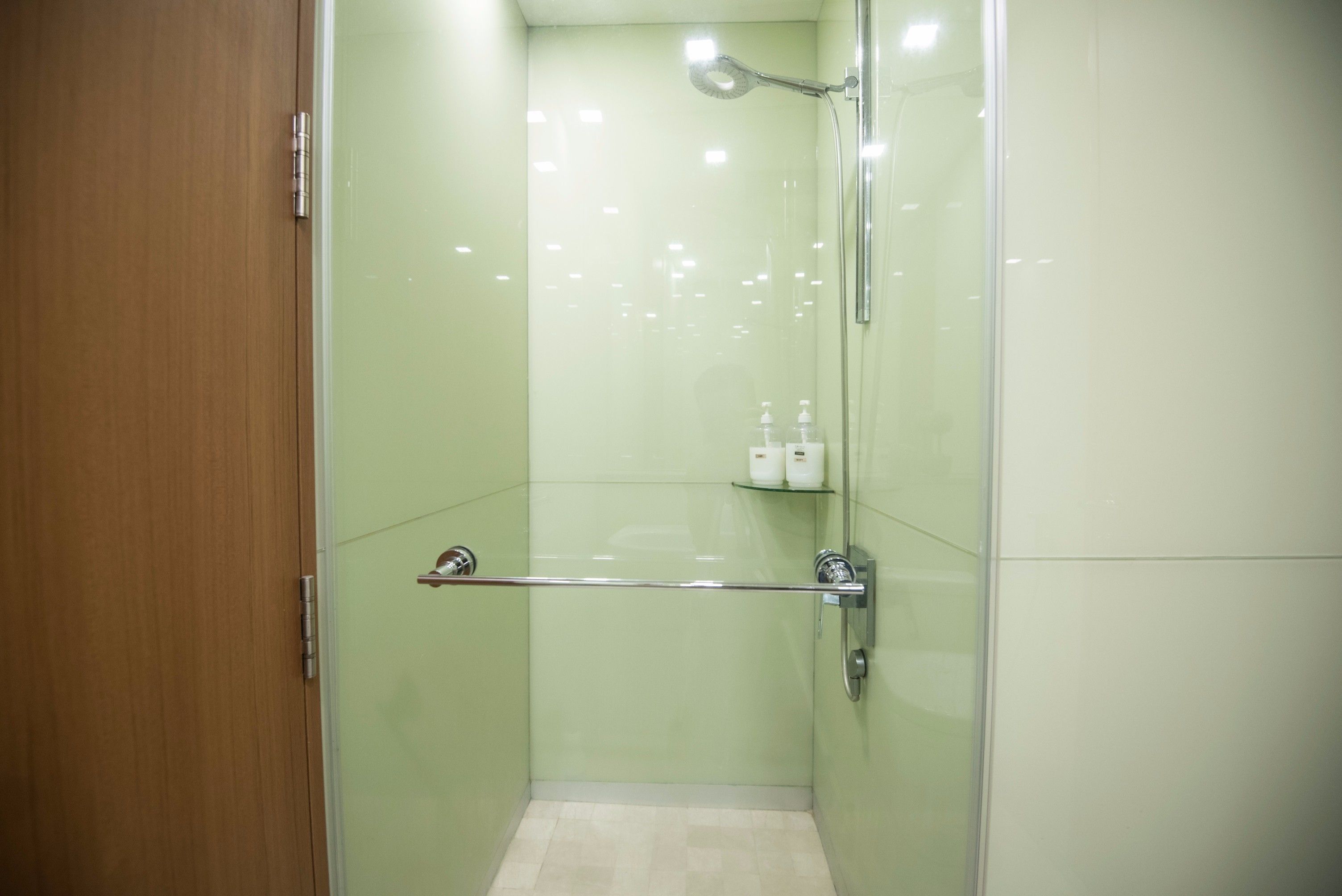 A glass-enclosed shower with a handheld showerhead, a glass shelf with toiletries, and a wooden door.