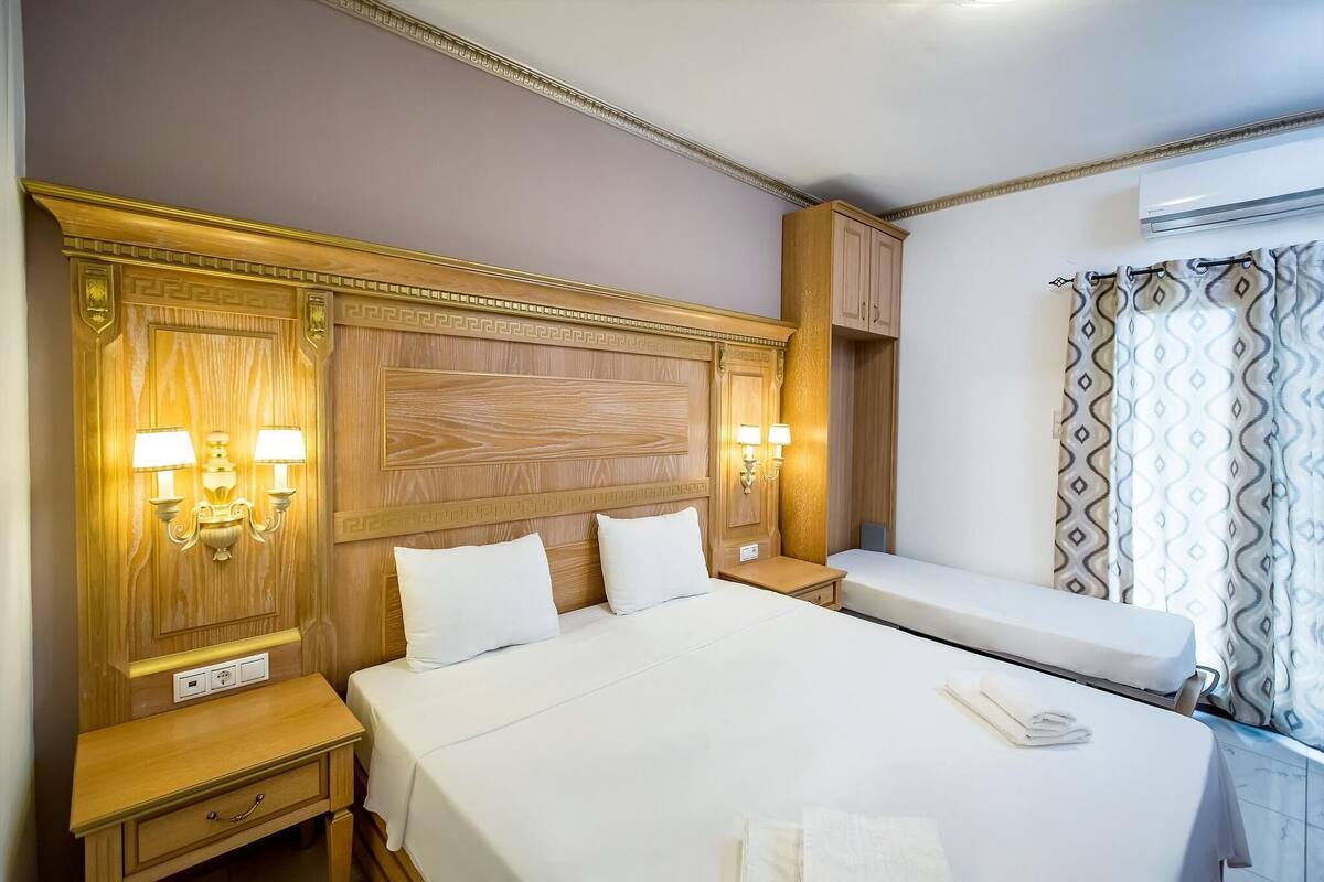 A hotel room with a large bed, two bedside tables, a nightstand, a wooden headboard, wall sconces, a window with patterned curtains, and an air conditioning unit.