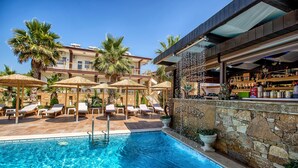 A swimming pool with lounge chairs and umbrellas, a bar area with a stone wall, and a building with balconies and palm trees in the background. - Greek Pride Ηotel and Apartments (Kassandra)