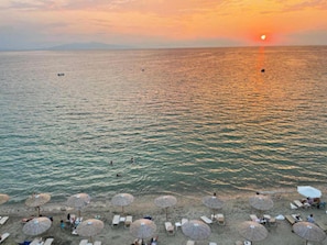 A beach with umbrellas and sun loungers, a sunset in the background, and a calm sea.
