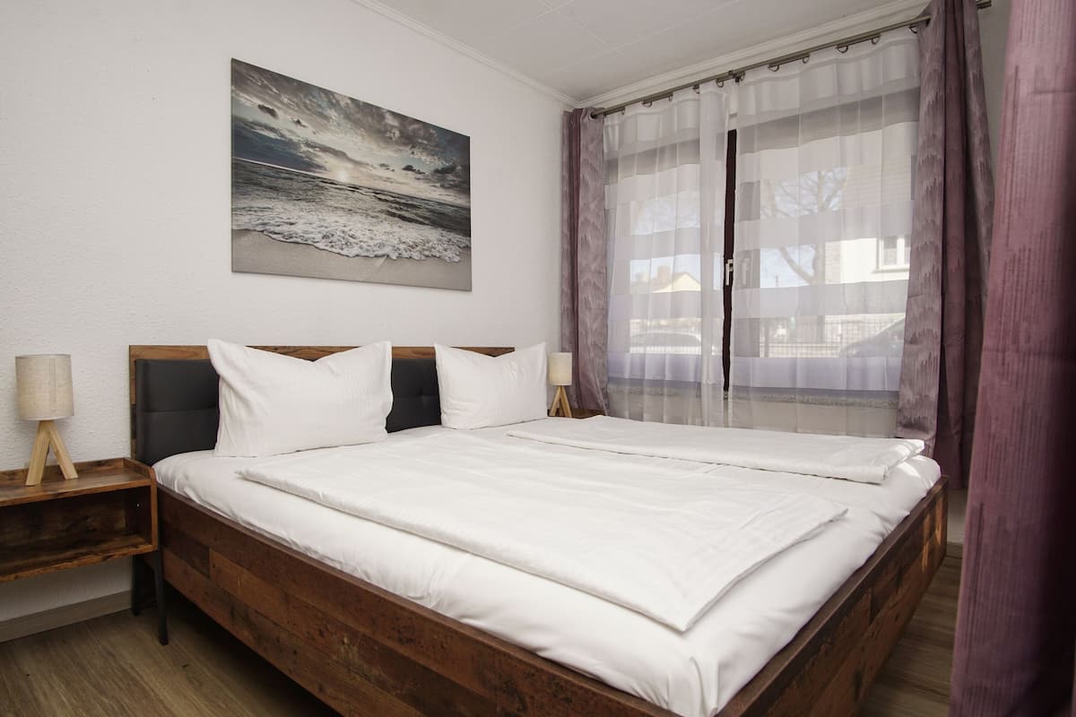 A double bed with white bedding, a wooden headboard, a bedside table with a lamp, and a painting of a beach scene on the wall.