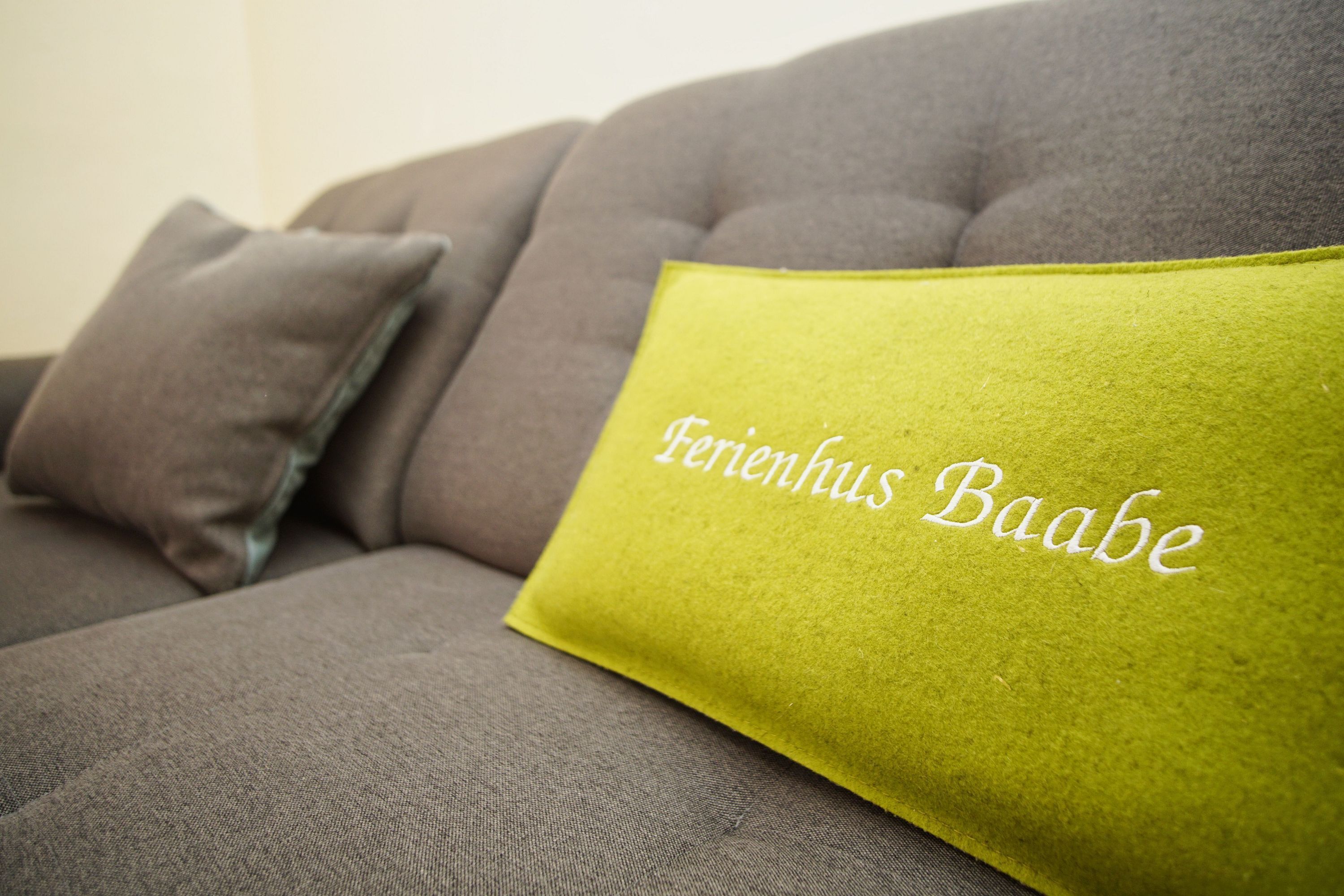 A grey sofa with a green cushion that has "Farenhus Baabe" embroidered on it.