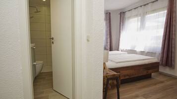 A bedroom with a wooden bed, a window with curtains, a shower area, and a white door.