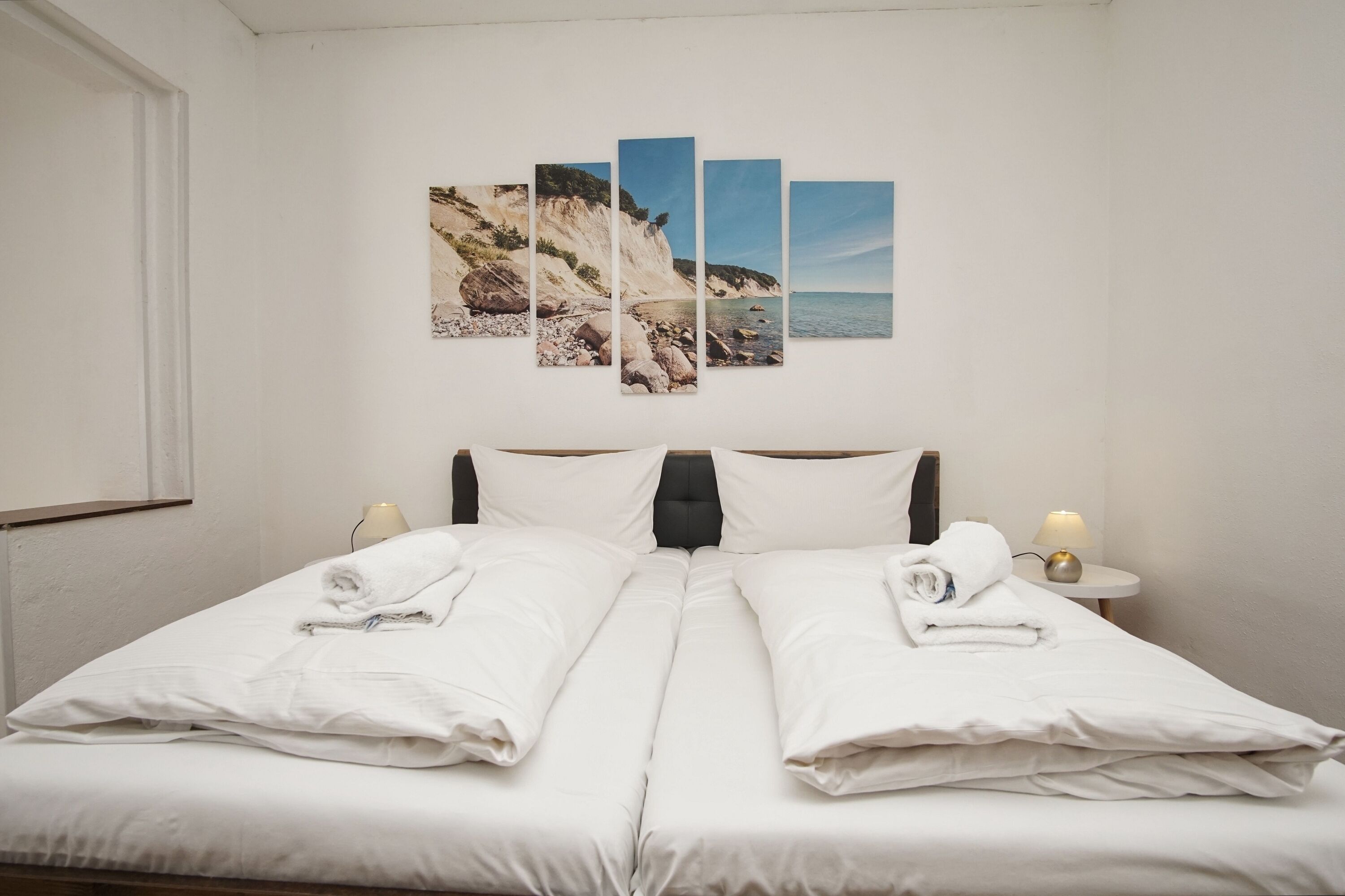 Ein Doppelbett mit weißen Leinen, ein Kopfteil, Nachttischlampen und ein an der Wand hängendes Kunstwerk mit einer Küstenansicht.