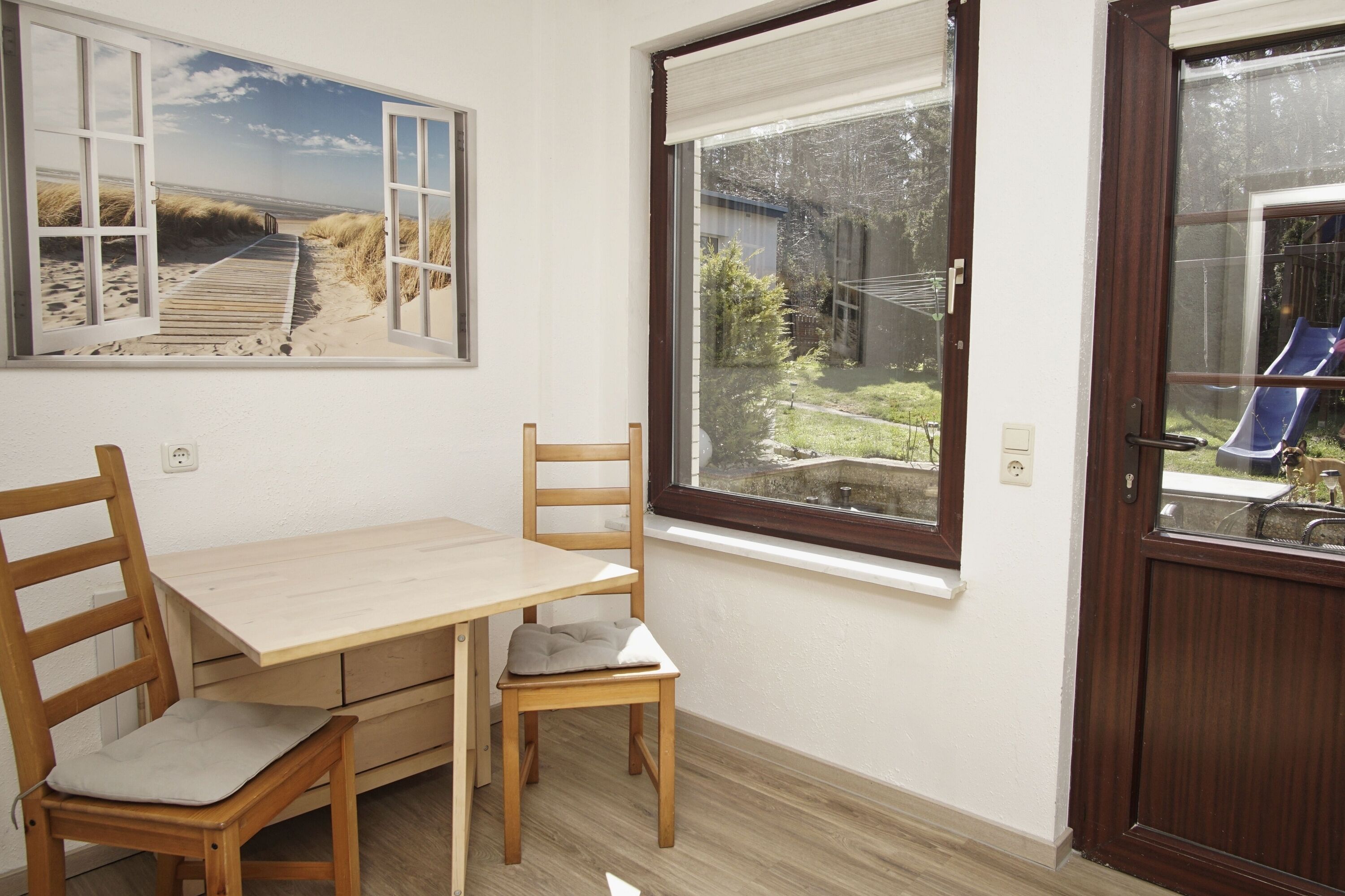A room with a wooden table and chairs, a window with a view of a garden and a slide, and a door with a window.