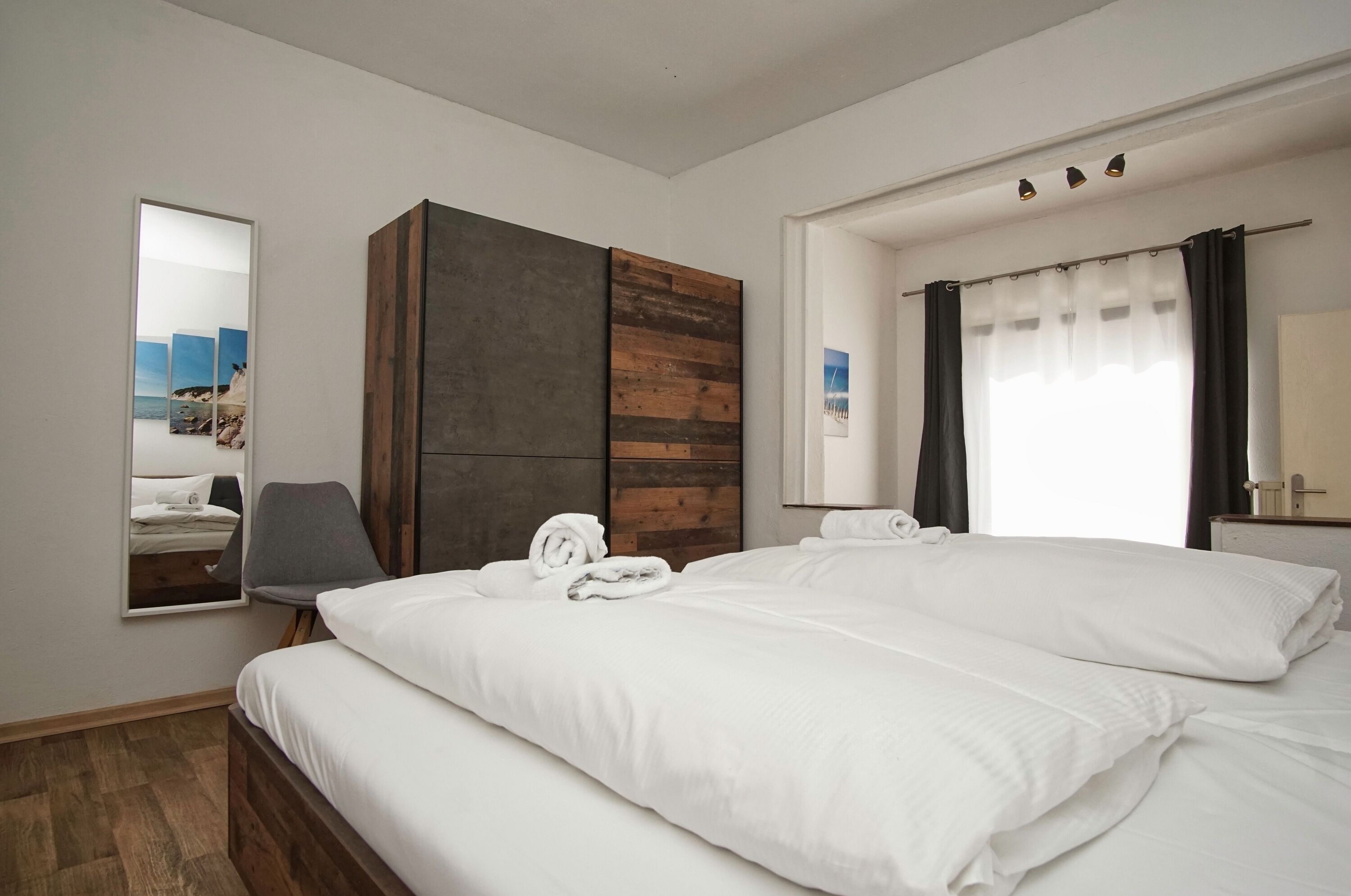 Habitación de hotel con cama, armario, silla, espejo y ventana con cortinas.