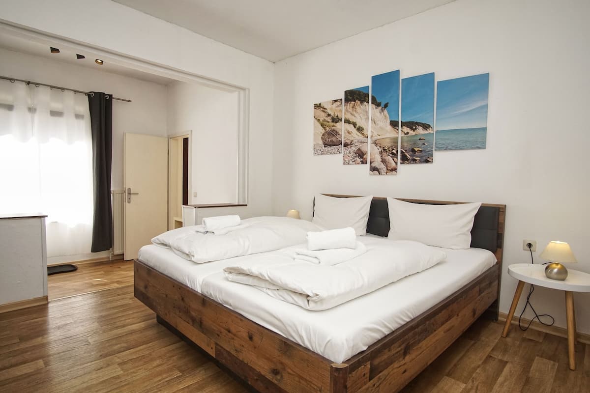 A bedroom with a wooden bed, white bedding, a bedside table with a lamp, and a wall with three landscape paintings.