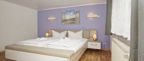 A neatly made bed with white linens and a beige headboard, a nightstand with a lamp, and a painting of a beach scene on the wall.