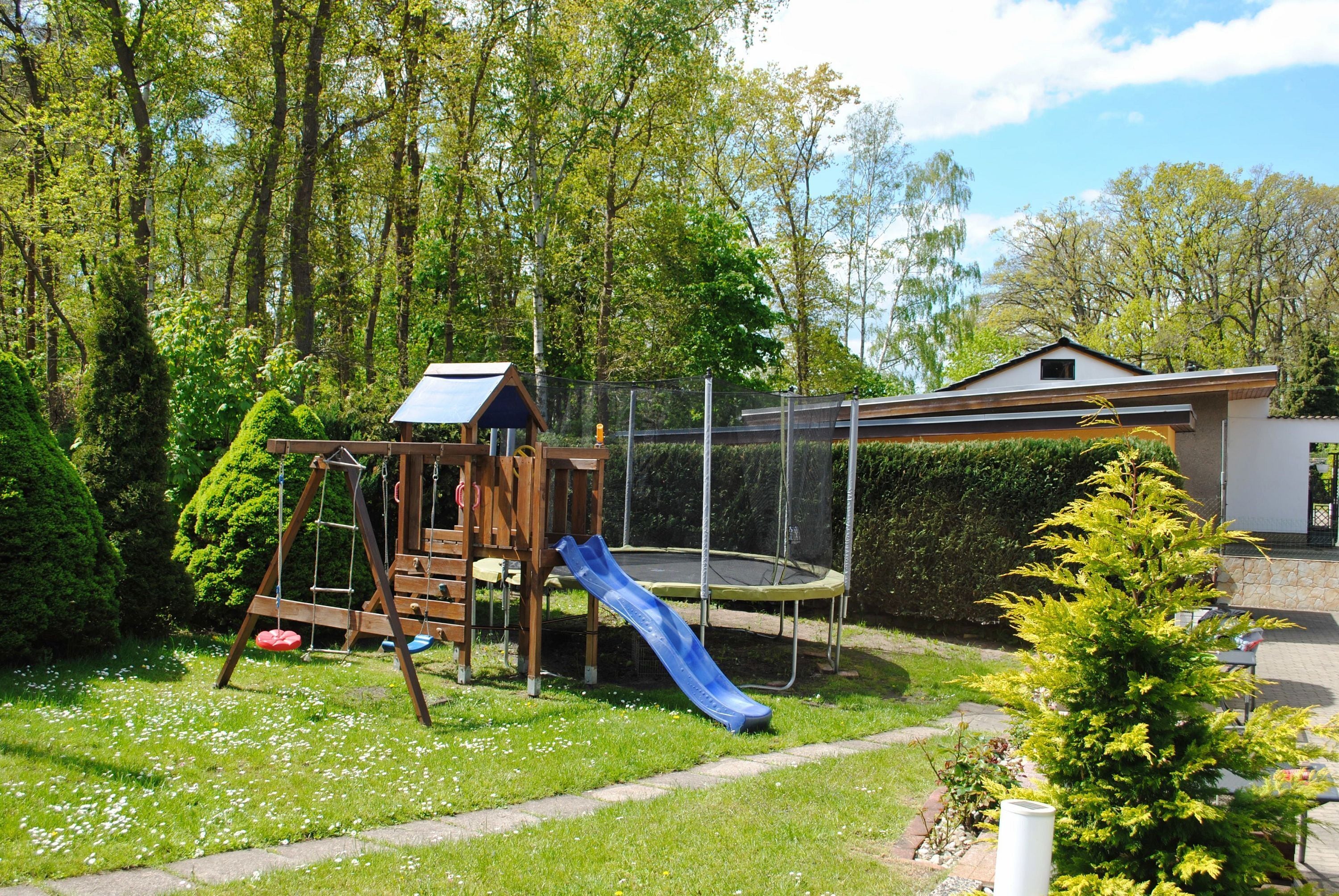 Een speeltuin met een glijbaan en schommels, omringd door bomen en een huis op de achtergrond.