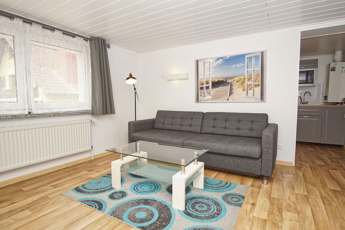 A modern living room with a glass coffee table, grey sofa, and a beach-themed painting.