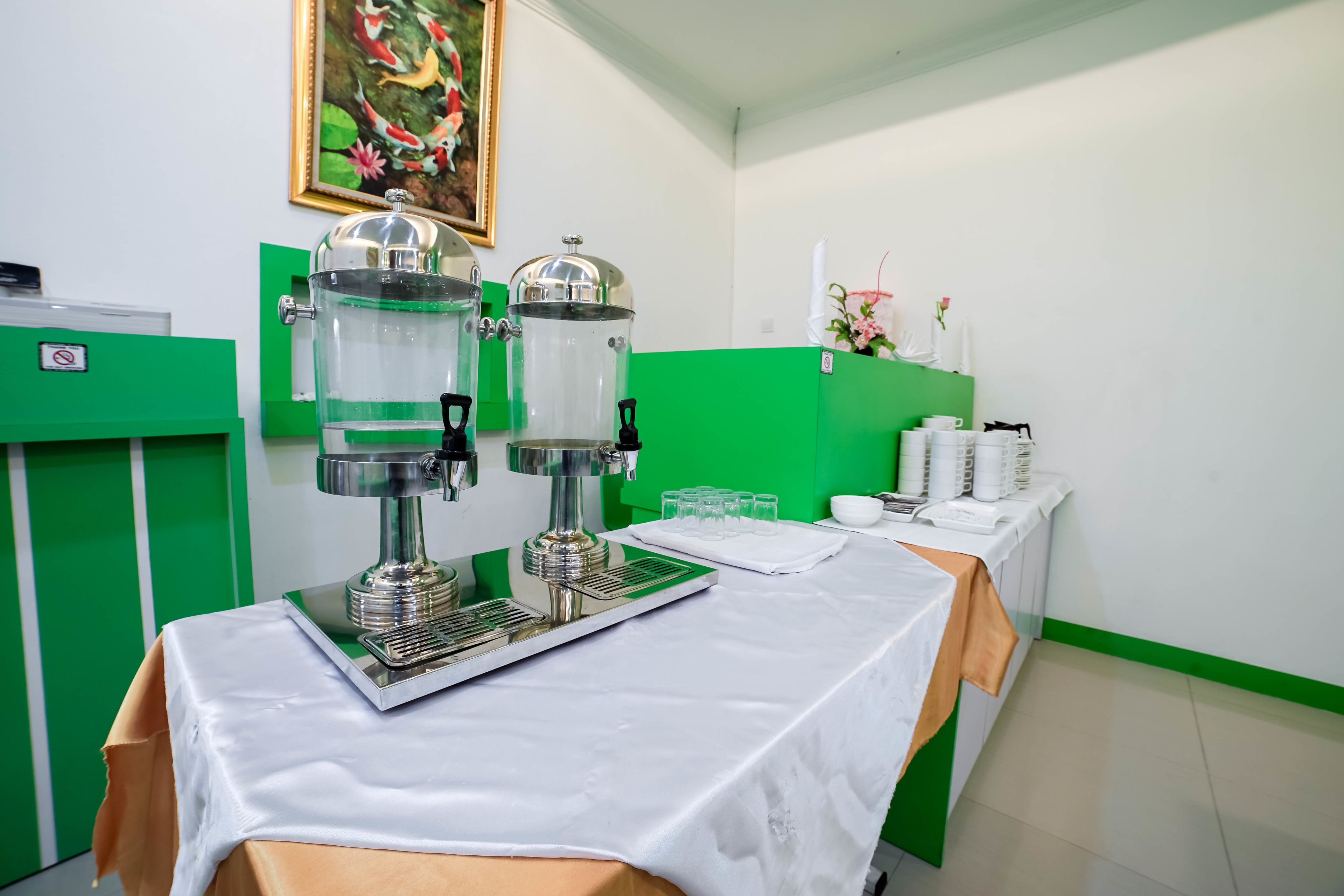 Habitación con una mesa dispuesta para una estación de bebidas, equipada con dos dispensadores grandes, una bandeja y un mantel blanco.