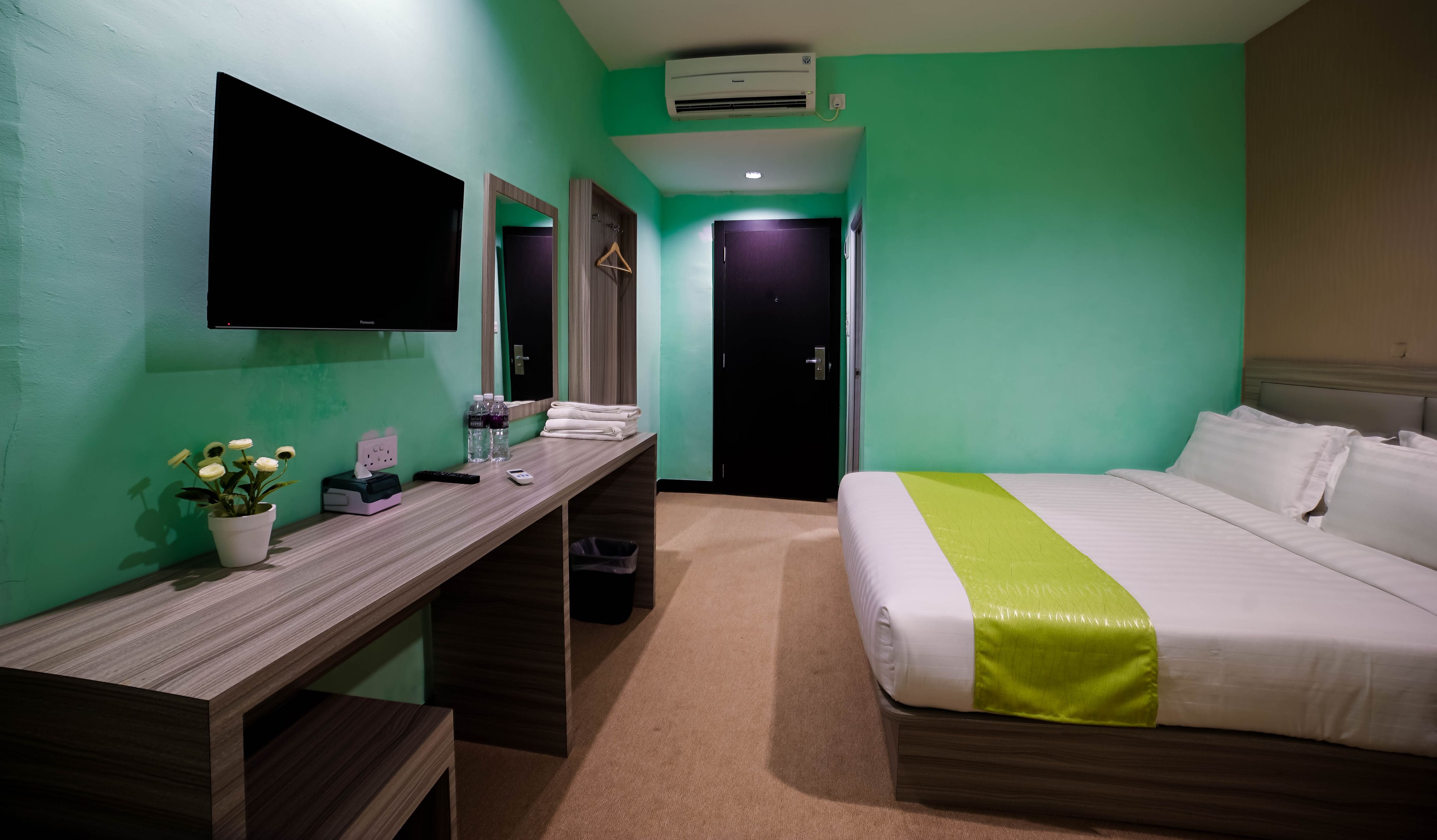 A hotel room with a bed, a desk with a flat-screen TV, a mirror, a wall-mounted air conditioner, and a door.