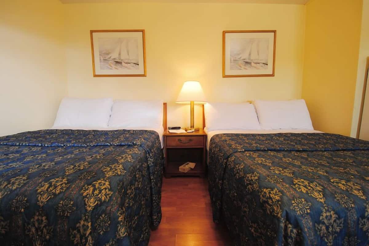 A hotel room with two beds, each with a blue patterned bedspread, a bedside table with a lamp, and two framed pictures on the wall.