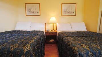A hotel room with two beds, each with a blue patterned bedspread, a bedside table with a lamp, and two framed pictures on the wall.
