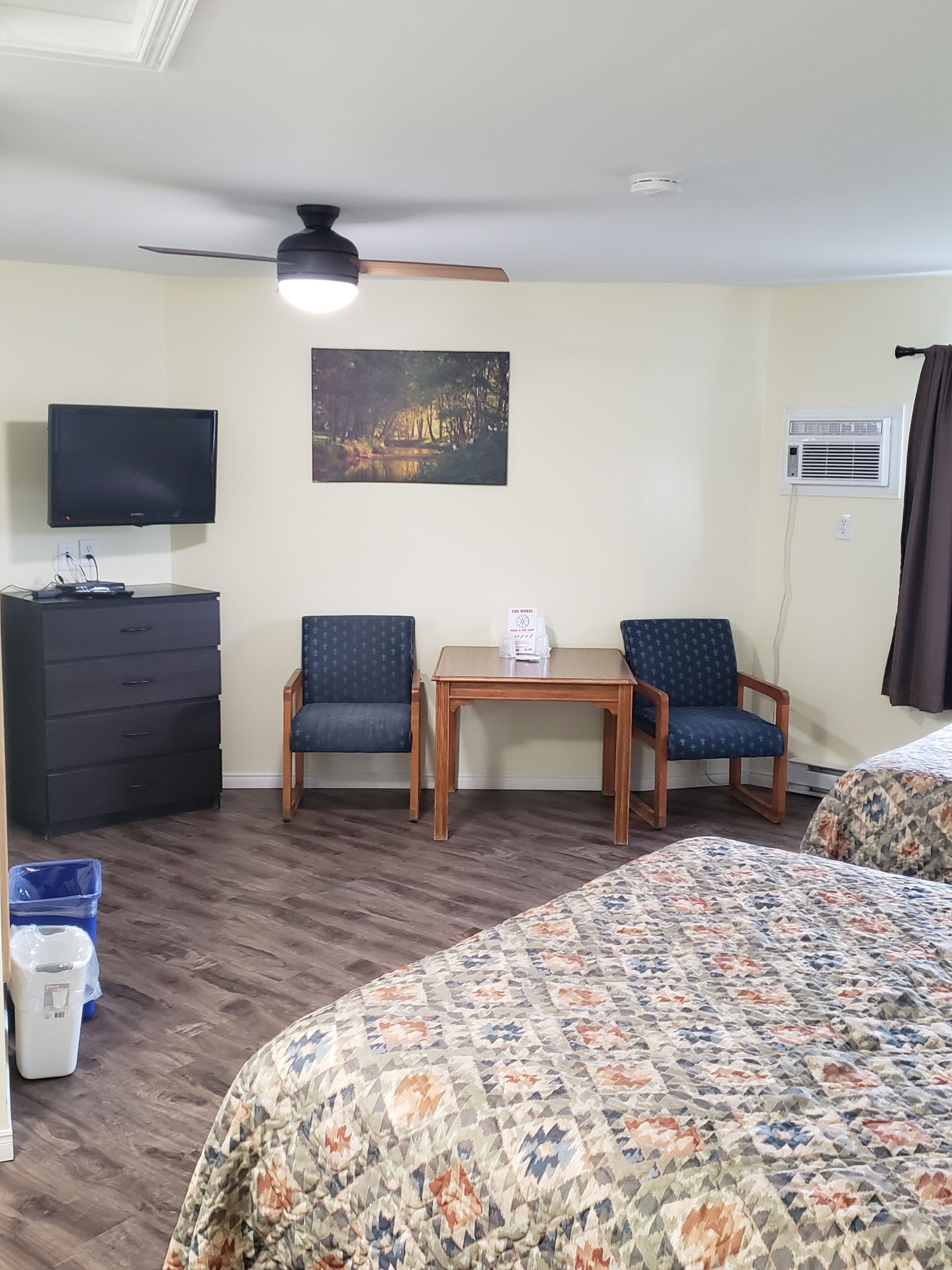 Una habitación de hotel con una cama, un escritorio con dos sillas, un televisor, un cuadro y un equipo de aire acondicionado.