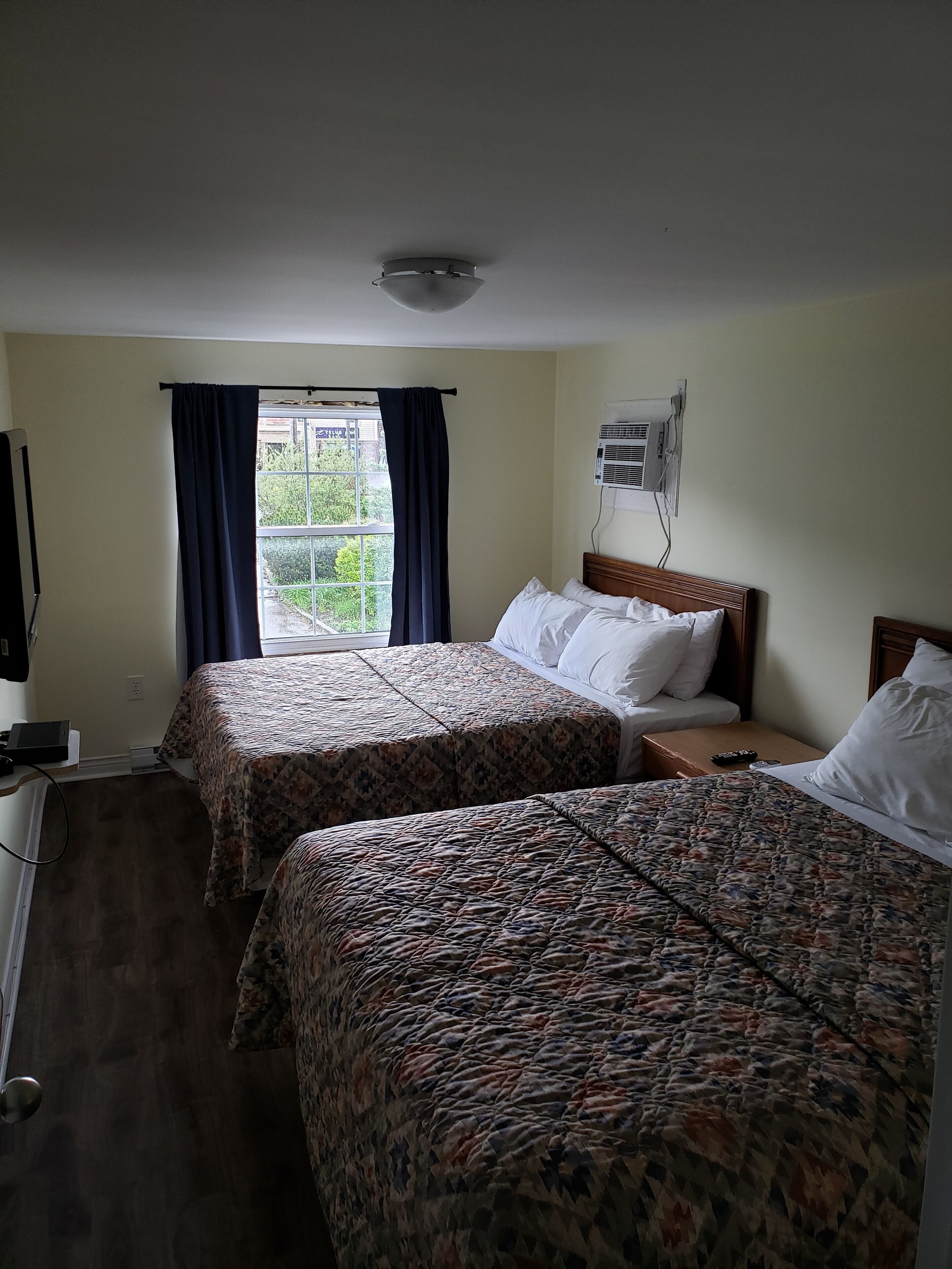 A hotel room with two beds, a window with curtains, an air conditioning unit, and a TV on the left.