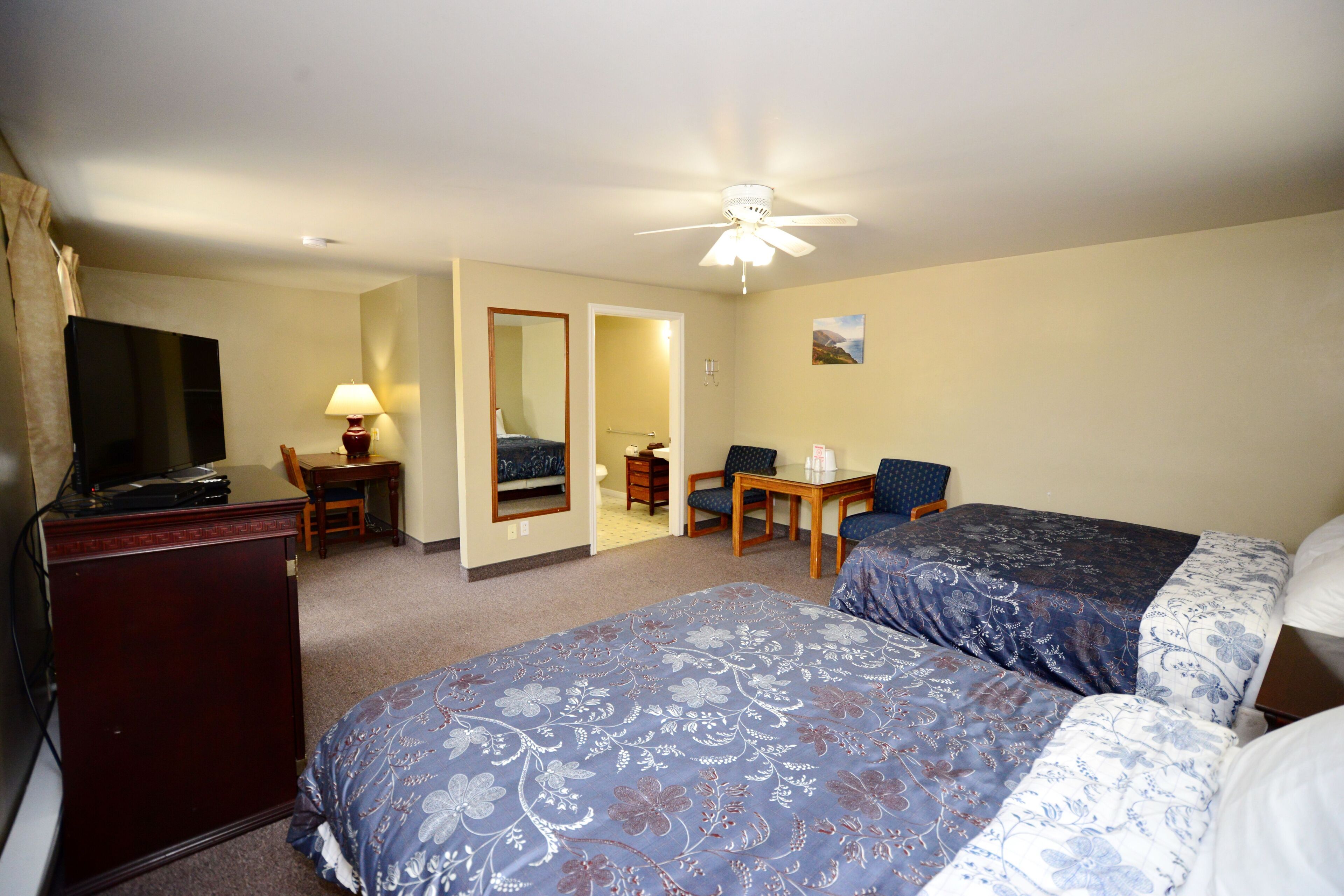 A hotel room with two beds, a desk, a chair, a TV, and a ceiling fan.