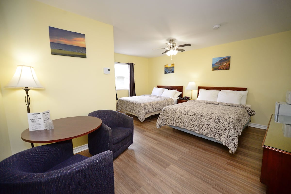 A hotel room with two beds, a ceiling fan, a desk with a chair, and a small table with a lamp.