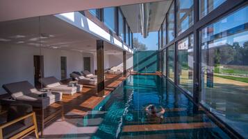 Una piscina interior moderna con paredes de vidrio, sillones de descanso y vistas al exterior.