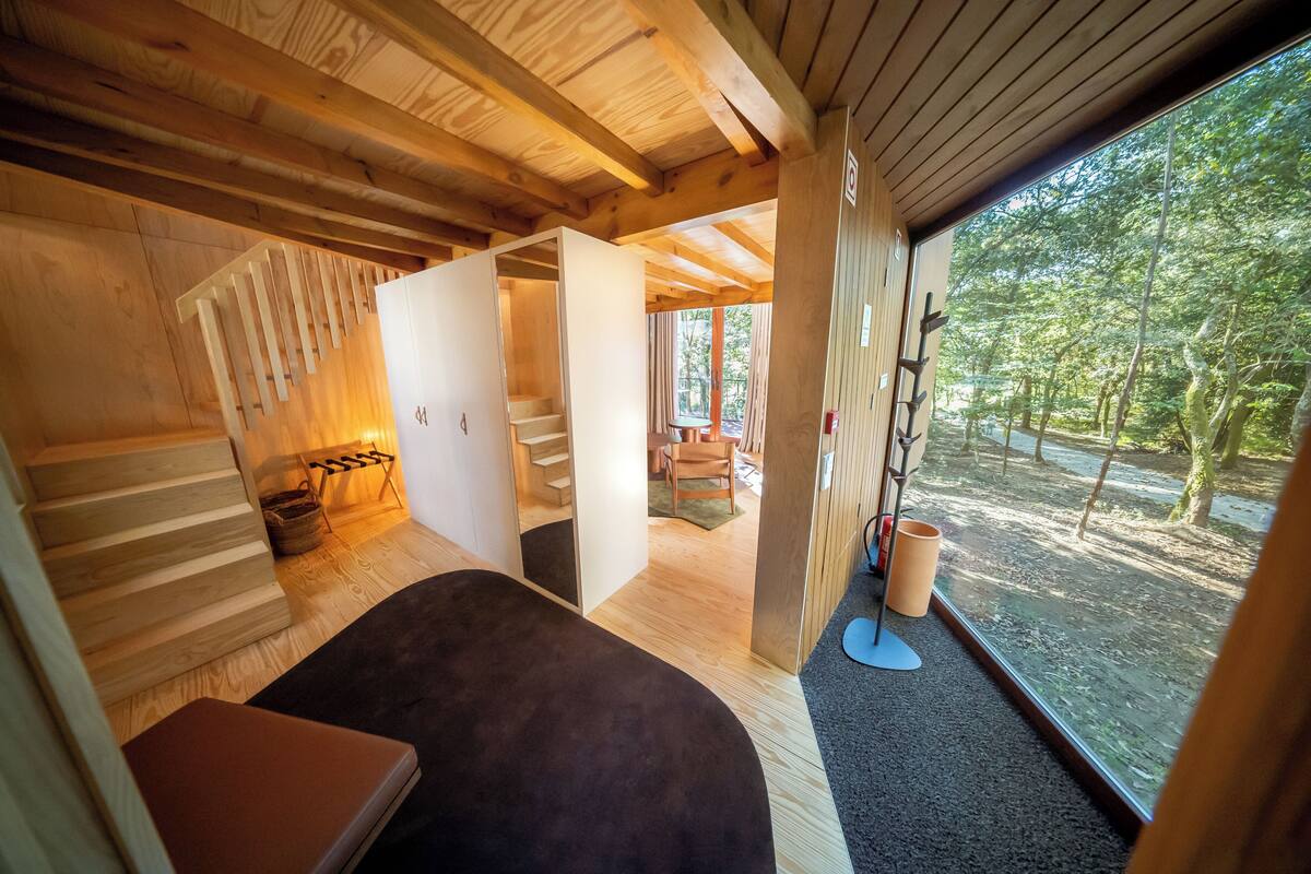 A wooden interior with a bed, a staircase, and a view of the forest.