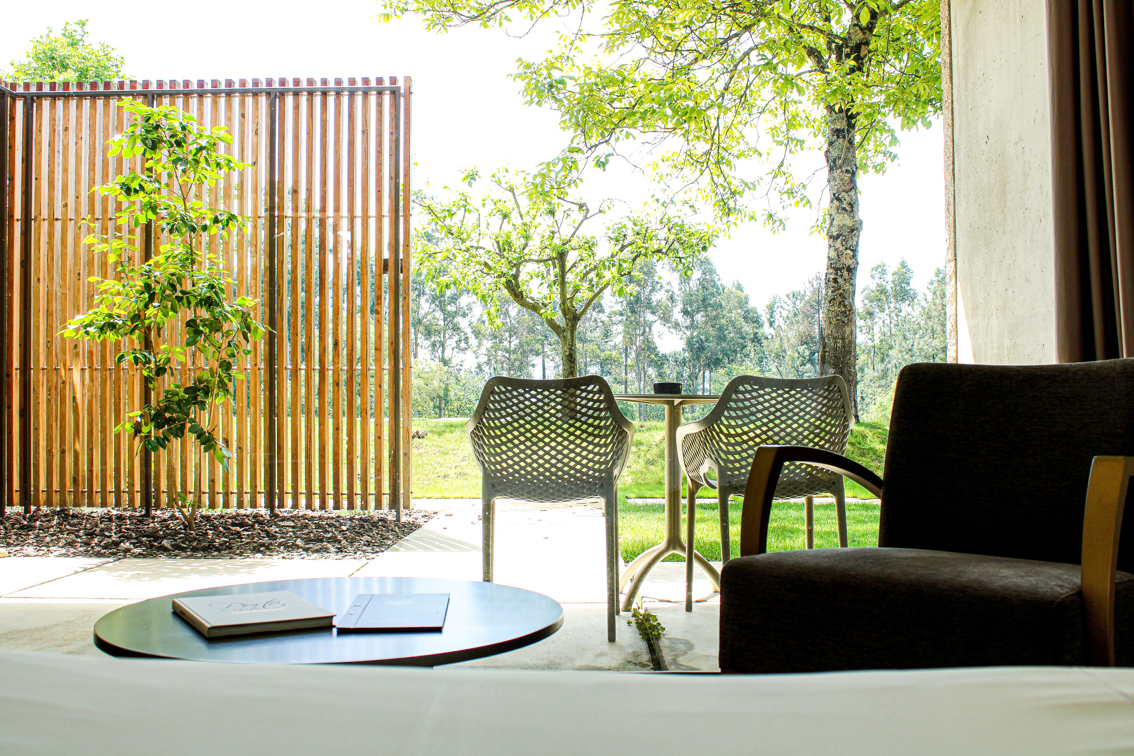 Una habitación con cama, una mesa redonda con un libro, dos sillas y vistas a un jardín y árboles a través de una ventana.