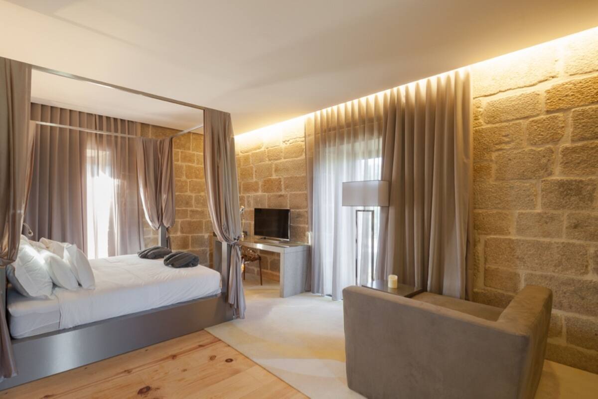 A modern bedroom with a large bed, a desk, and a chair. The room has stone walls and a wooden floor.