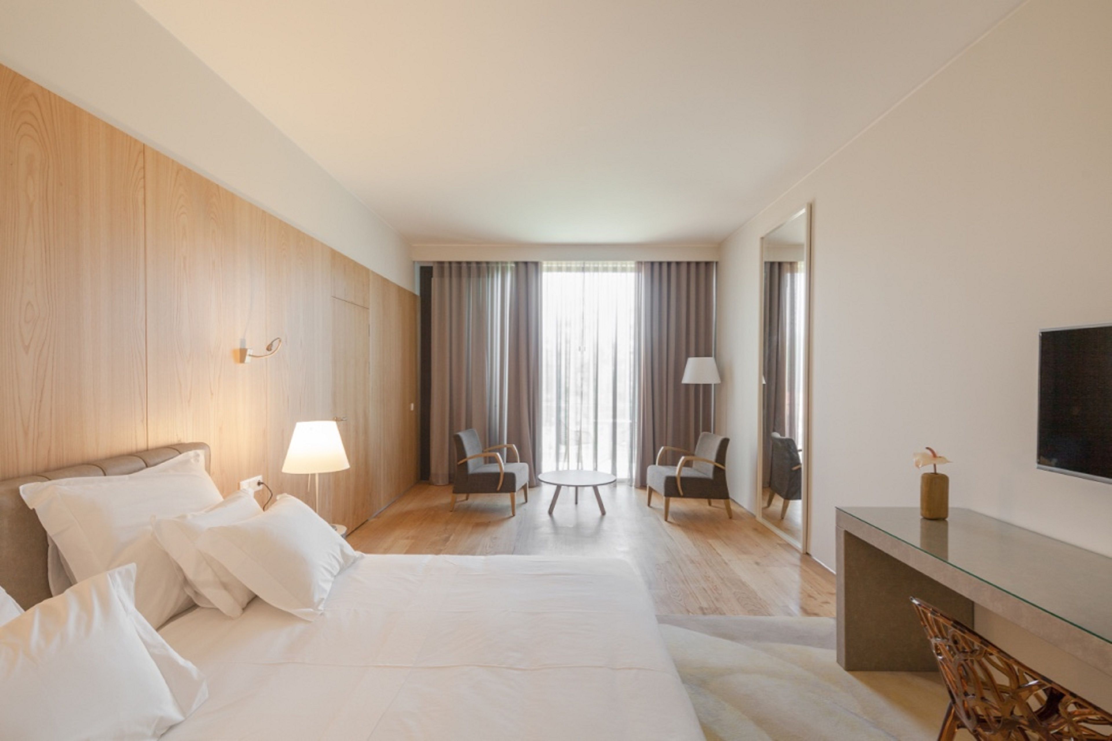 A modern hotel room with a large bed, two armchairs, a small table, a desk, and a TV.