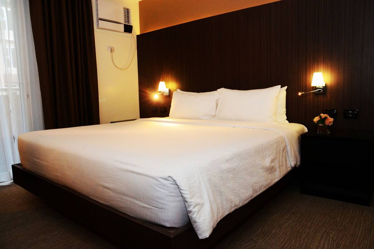 A hotel room with a large bed, two bedside lamps, a nightstand with a vase of flowers, and a wall-mounted air conditioning unit.