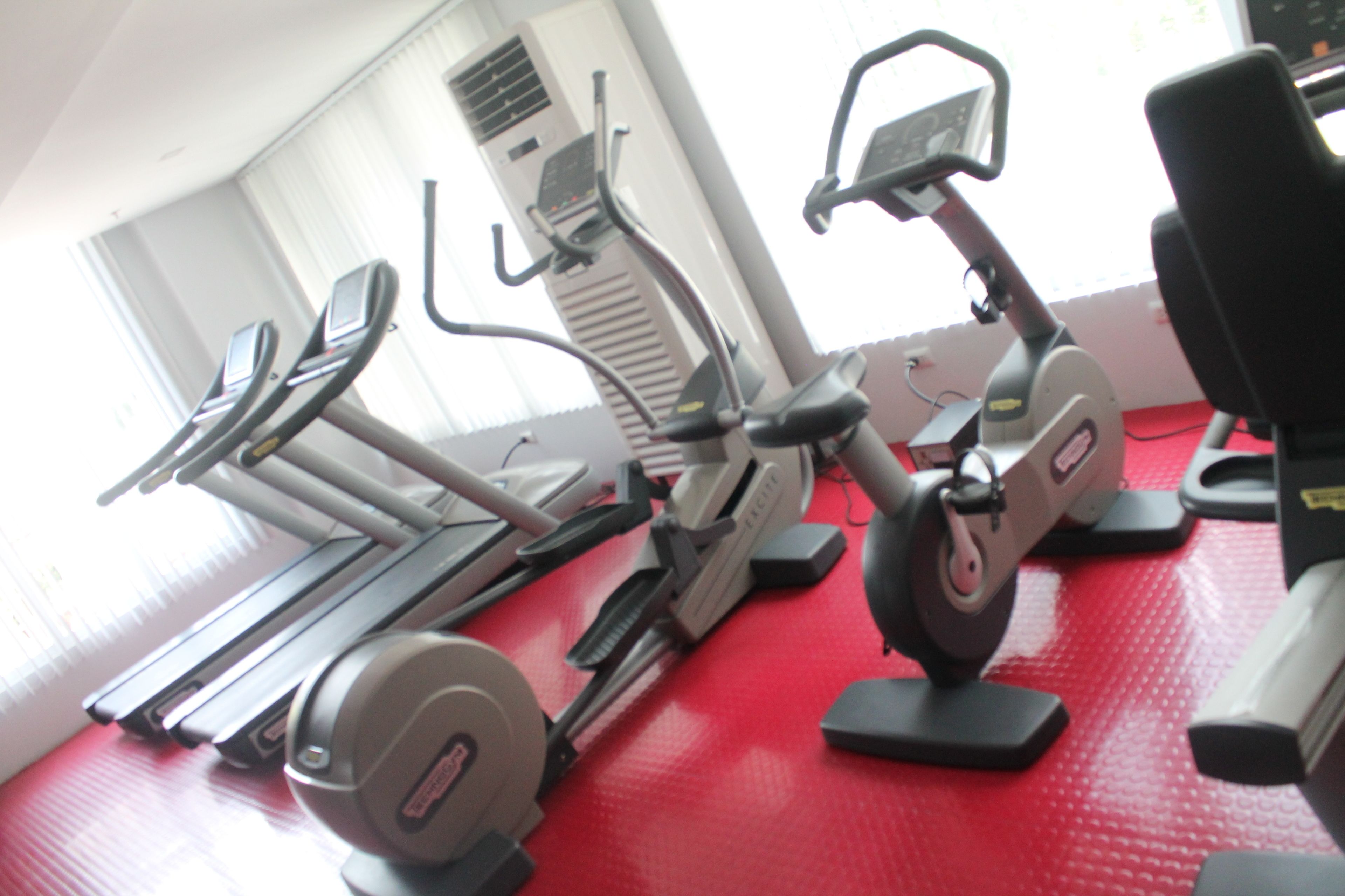 A gym with various exercise machines, including treadmills, elliptical trainers, and stationary bikes, arranged on a red floor.