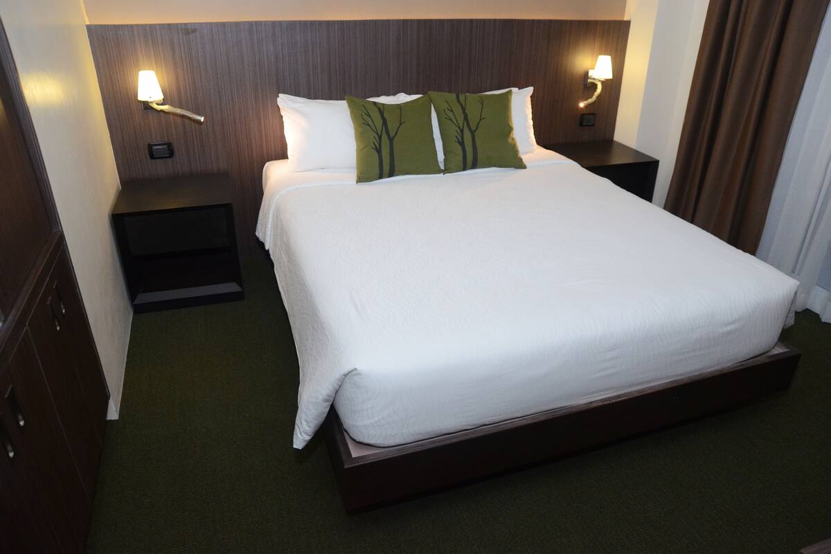 A neatly made bed with white linens and two green tree-patterned pillows, flanked by two bedside tables with lamps, in a room with dark wood paneling and a green carpet.